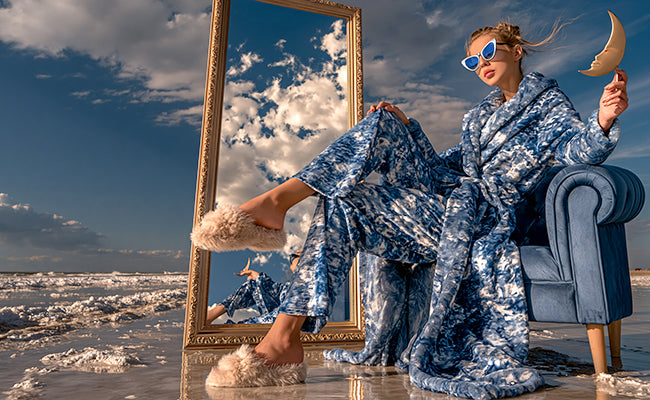 Selbstbewusste Frau mit Sonnenbrille auf einem blauen Sessel am Strand neben einem grossen, goldfarbenen Spiegel.