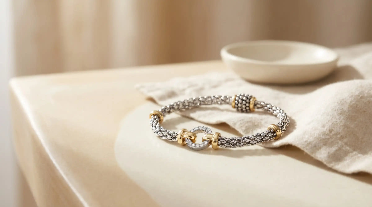 Silberfarbenes Armband mit Magnetverschluss und goldfarbenen Akzenten auf beigem Tisch neben Leinentuch. Avirelle.