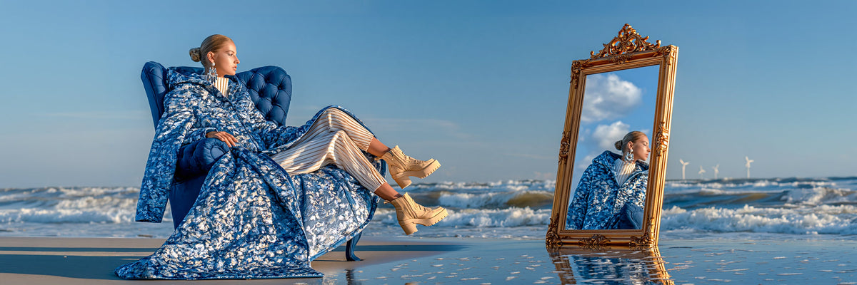 Selbstbewusste Frau auf einem grossen blauen Sessel am Strand neben einem prunkvollen goldfarbenen Spiegel, der sie reflektiert, während sie auffällige silberfarbene Ohrringe trägt.