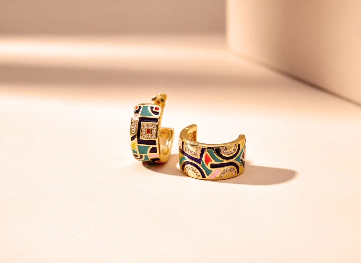Goldfarbene, halboffene Creolen mit geometrischem Emaille-Muster und feinen Ziersteinen auf warmem Hintergrund.