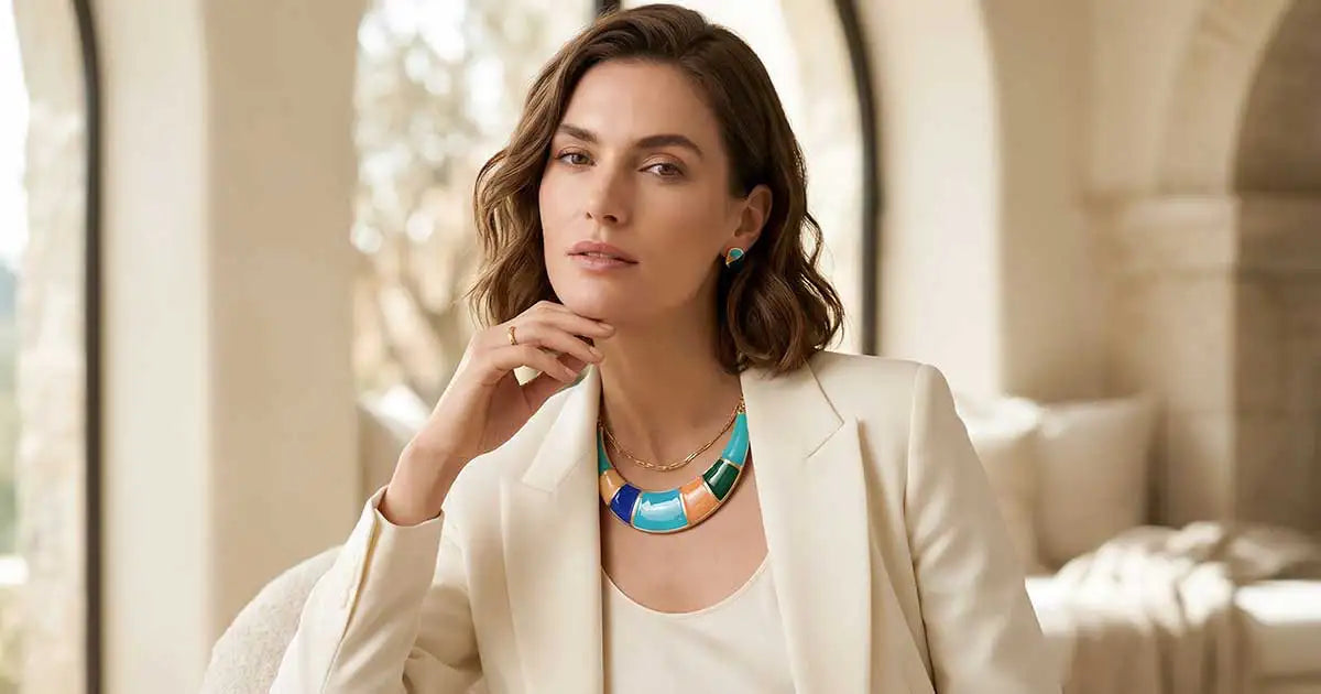 Selbstbewusste Frau im schwarzen Off-Shoulder-Top mit markantem goldfarbenem Schmuck – die perfekte dunkle Basis, um farbigen Emaille Schmuck kombinieren zu können.