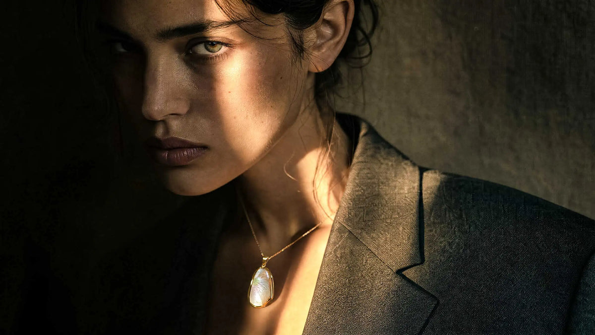 Nahaufnahme einer souveränen Frau mit dunklem, strukturiertem Blazer, die ein modernes, roségoldfarbenes Collier mit einem grossen, ovalen Perlmutt-Anhänger trägt. Das Material schimmert im dramatischen Licht-Schatten-Spiel.