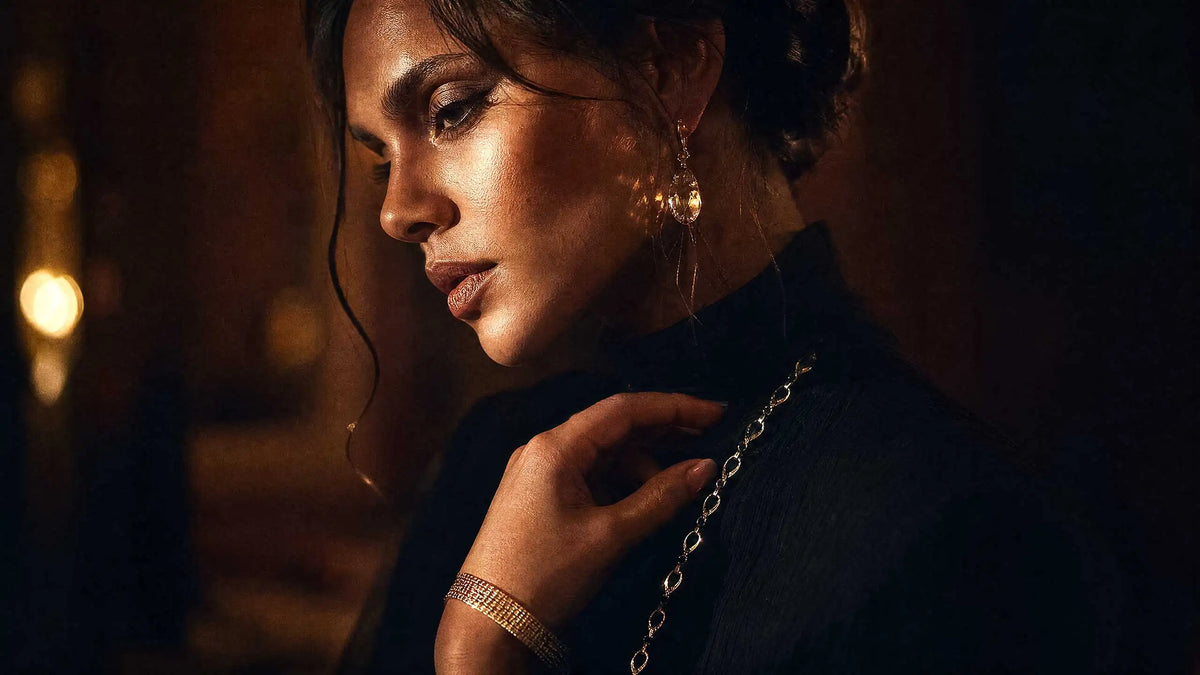 Elegante Frau in schwarzem Abendoutfit kombiniert mit auffälligen, mehrlagigen goldfarbenen Halsketten, passenden Ohrringen und Armbändern vor unscharfem Hintergrund mit Abendbeleuchtung.